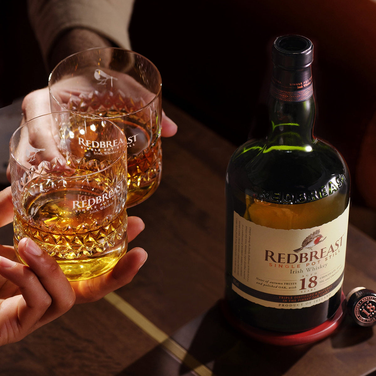 Schwarzbach Spirits Redbreast 18 Year Old Irish Whiskey bottle next to two hands holding filled crystal glasses